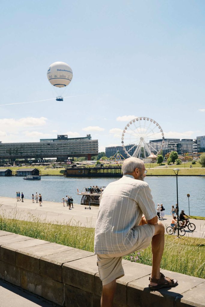 Older man resting one leg on a stone wall, looking at the Vistula River in Krakow, with the Forum Hotel, a Ferris wheel, and a hot air balloon in the background.