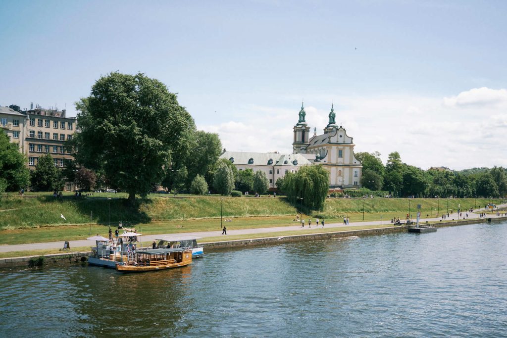 View of the Vistula riverbank in Krakow on a sunny day, featuring a floating boat cafe, people walking, and the historic Church on the Rock with twin green spires.