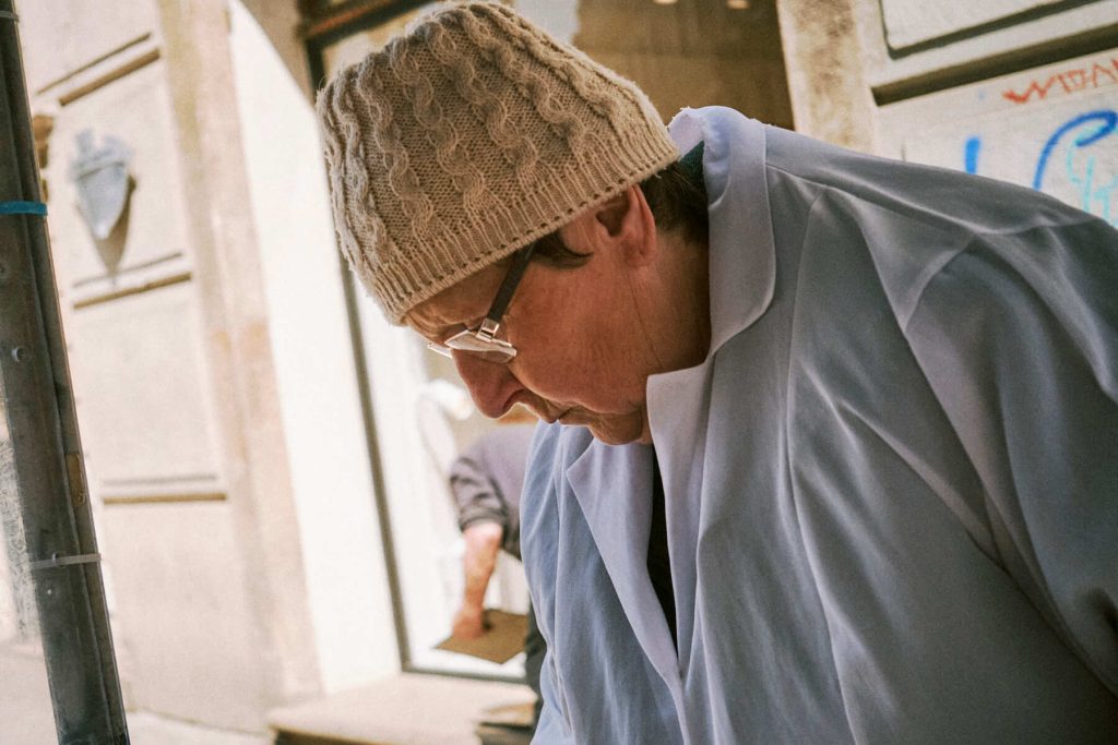 Close-up street portrait of an older woman wearing a beige knitted beanie, glasses, and a light blue coat, looking down.