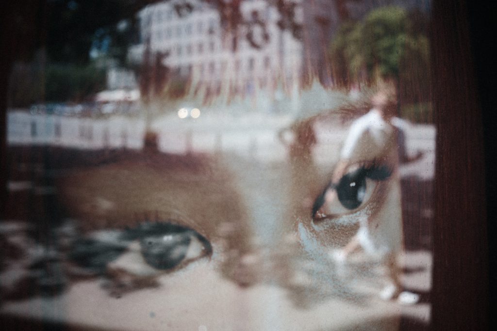 A close-up of a child's face seen through a reflective window, with a large eye in focus and blurred city buildings and street lights reflected across the glass.