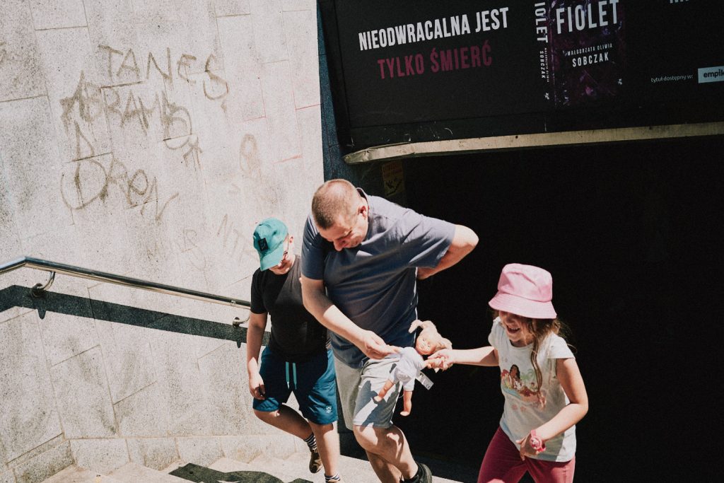 A family with two children walking up a set of stairs from an underground entrance, with one child holding a doll and bright sunlight casting strong shadows on the wall.