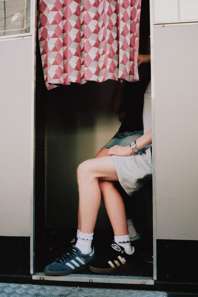 A person sitting inside a small fitting room, partially hidden behind a patterned curtain, with their legs, crossed hands, and Adidas sneakers visible.