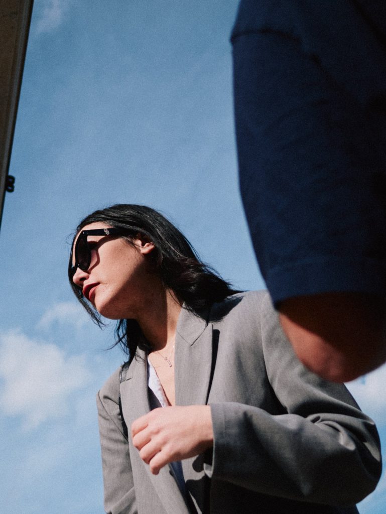 Candid street portrait of a woman in a grey blazer and sunglasses looking away against a blue sky