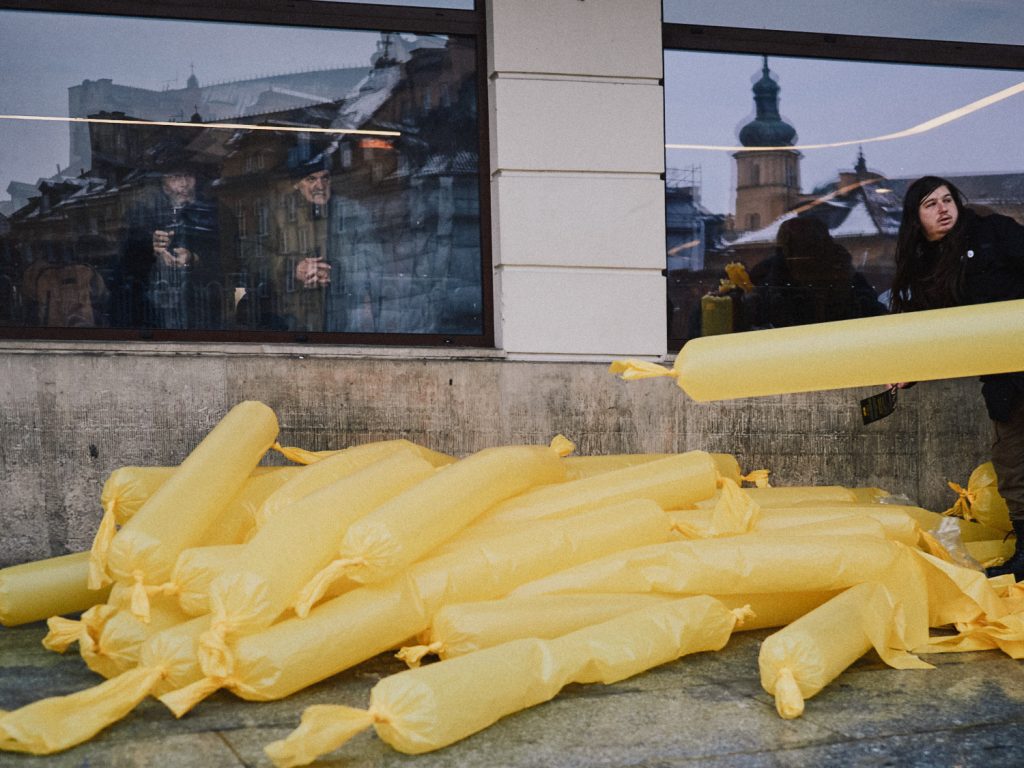A pile of bright yellow plastic-wrapped tubes on a city sidewalk, with people reflected in a window and historic buildings visible in the background.